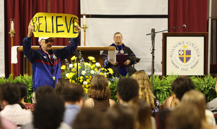 Chaplain Bowen, dressed as the character Ted Lasso, holds up a large sign bearing the handwritten word: BELIEVE