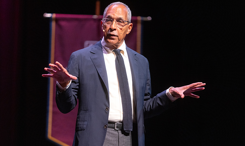Claude Steele, in a navy coat and tie, gestures with his hands as he talks on the Olin Theater stage.