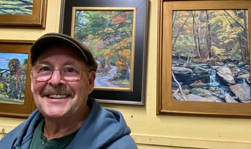 Man in a blue sweatshirt and brown tweed cap poses in front of a yellow wall where several landscape paintings are hung, including one of a stream running through the forest.