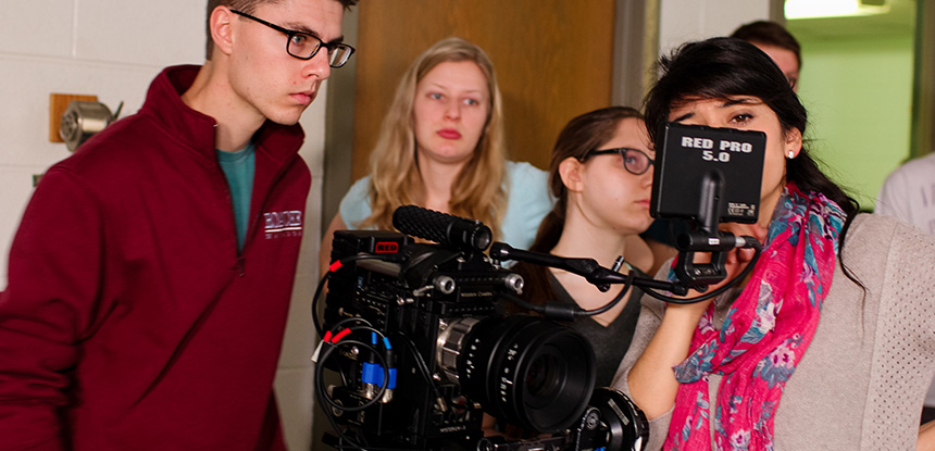 Students stand behind a video camera and look at the screen on it. 