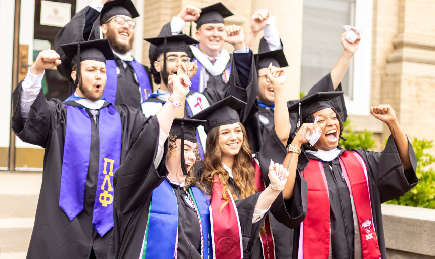Students in commencement robes cheer while raising their arms in the air