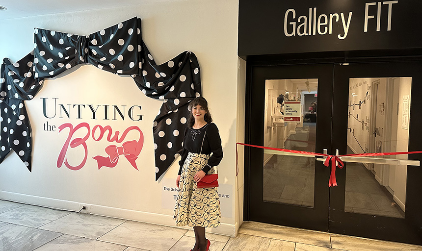 Woman in black-and-white outfit with red accessories stands outside Gallery FIT. There is a red ribbon across the entrance and a sign for the exhibition, "Untying the Bow" on the wall.