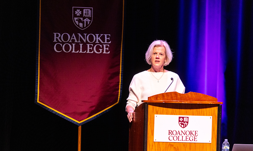 Woman with white hair stands behind a podium with a maroon Roanoke College banner hanging to her right.