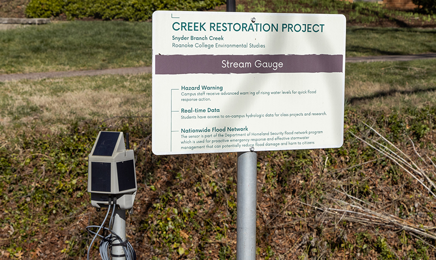 A sign along Snyder Branch Creek explains the flood sensor and creek restoration projects. To the left of the sign is one of the flood sensors, which looks like a gray box with solar panels mounted on a pole next to the creek.