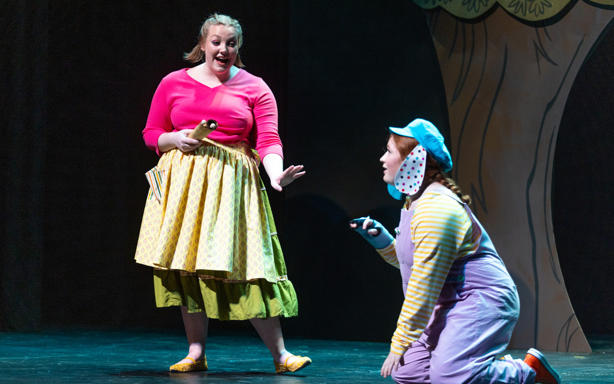 Two actresses, one dressed as a dog and kneeling on the ground, perform on stage