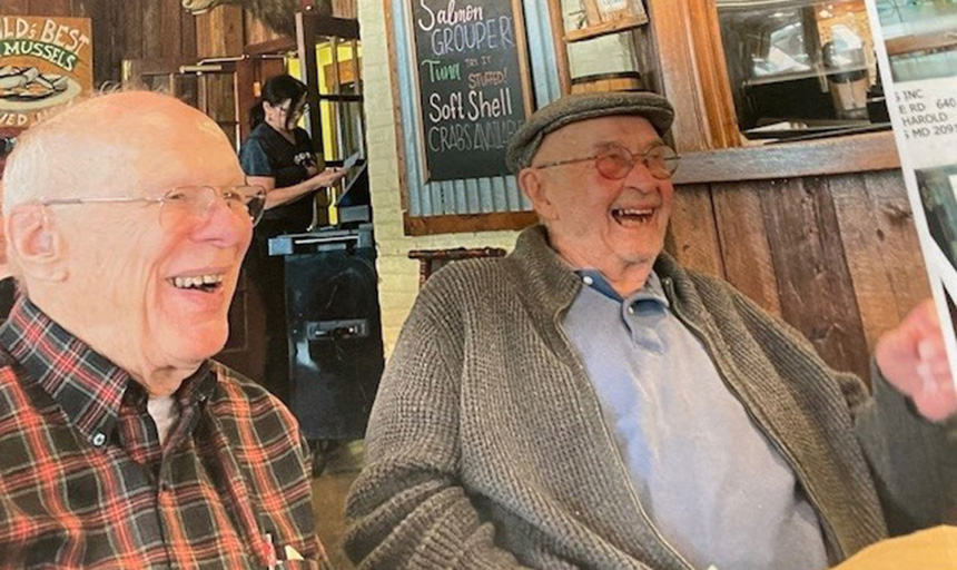 Two senior men sit at a table in a restaurant, laughing. In the background is a chalk board with seafood dishes written on it, including soft shell crabs, salmon and grouper.