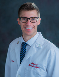 Headshot of a young, smiling man in dark-rimmed glasses and wearing a white doctor's coat with the words "Harvard Medical School" stitched in red.