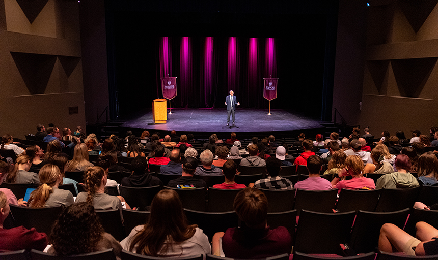 Photo taken from the back of a crowded room, with a man on stage under the lights. 