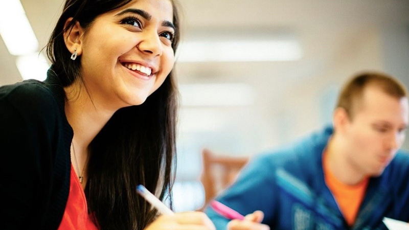 smiling student, seated at a table, writing with pen and paper