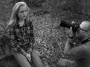 A man taking a picture of a girl sitting on a log in the forest