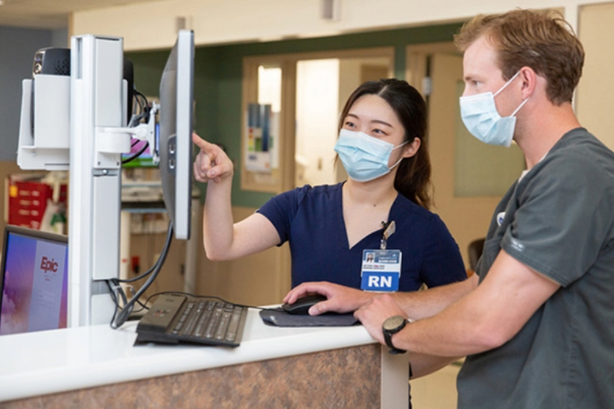Two health care providers wearing surgical masks discuss information displayed on a screen
