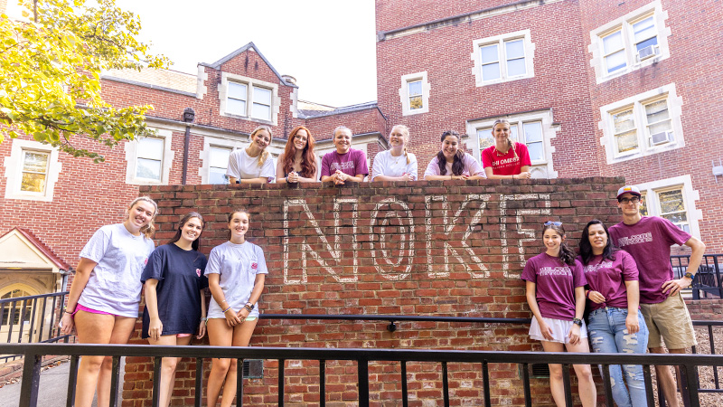 Students smiling next to the word Noke written in chalk outside a residence hall