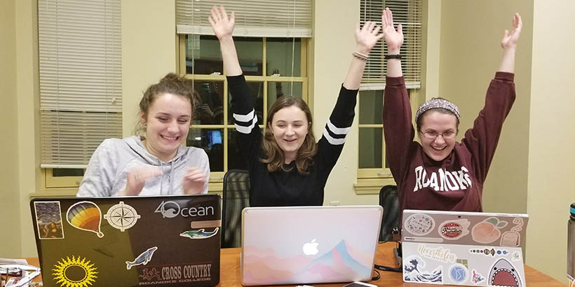 Three students with laptops. All are smiling, two have their hands up in the air