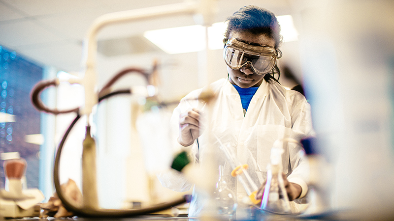 Lydiah Mpyisi works in a laboratory wearing a white coat and goggles.