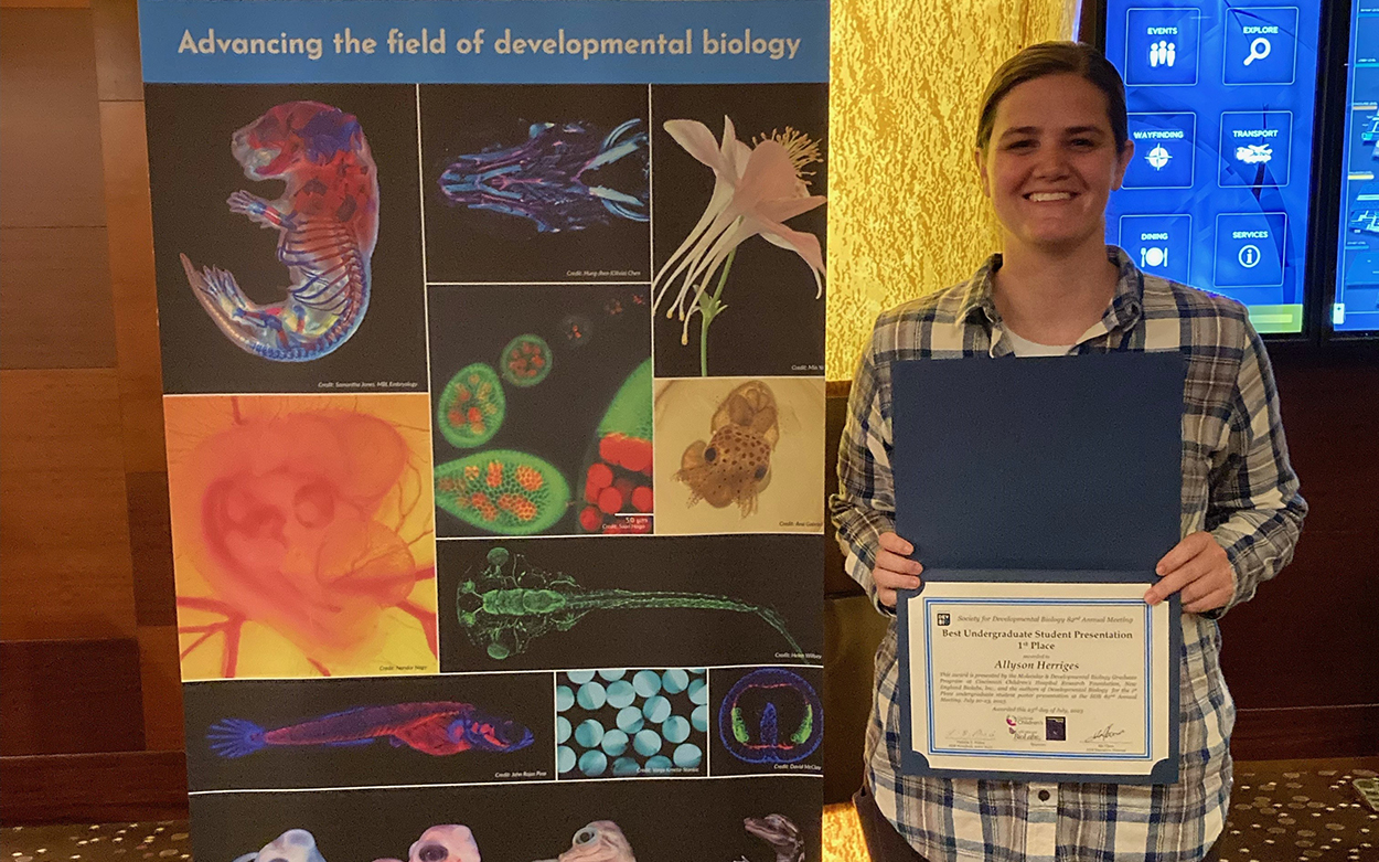 Allyson Herriges stands holding her certificate beside a poster for the Society for Developmental Biology's 82nd annual meeting 
