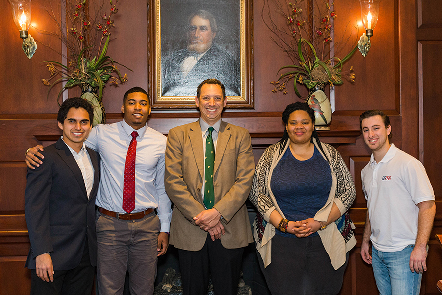 A man in a tan suit stands in the center with two students on each side. They are in a wood-paneled room and smiling at the camera.