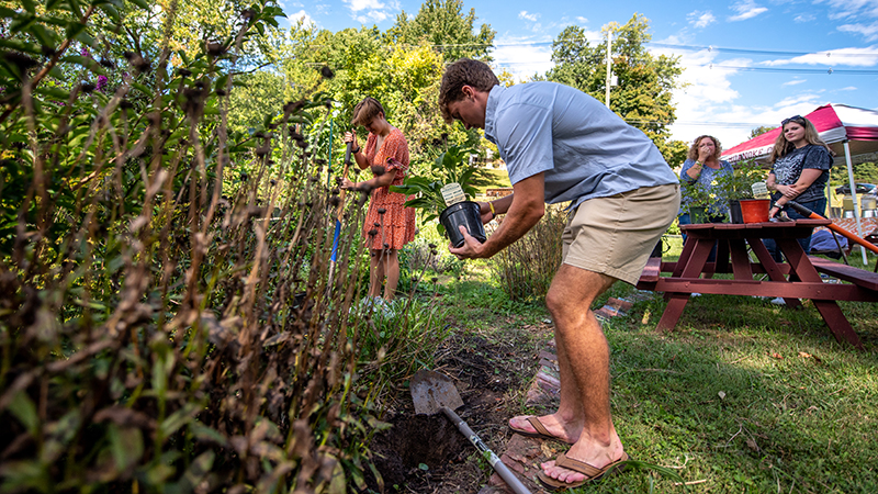 A student works plants a plant in the campus garden while others look on under a brilliant blue sky.