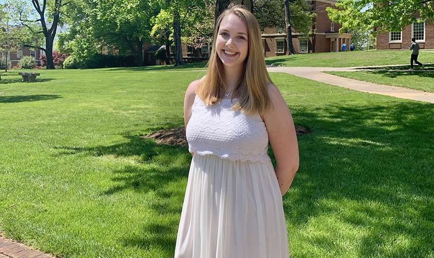 student in white dress stands with a lawn behind her