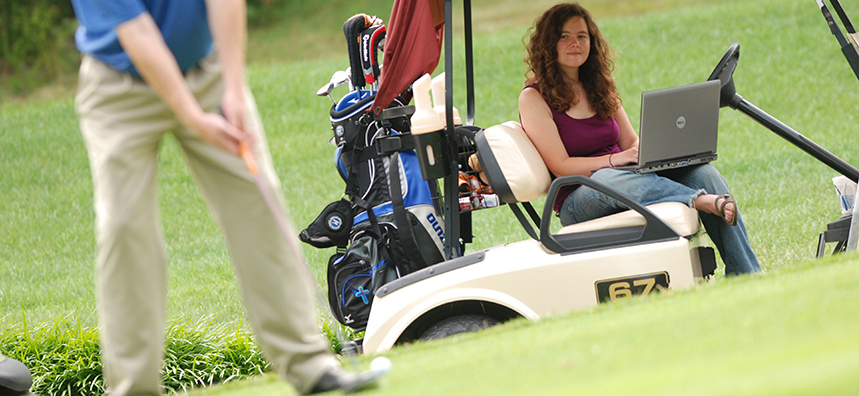 woman sitting in a golf cart holding a laptop while she observes someone playing golf