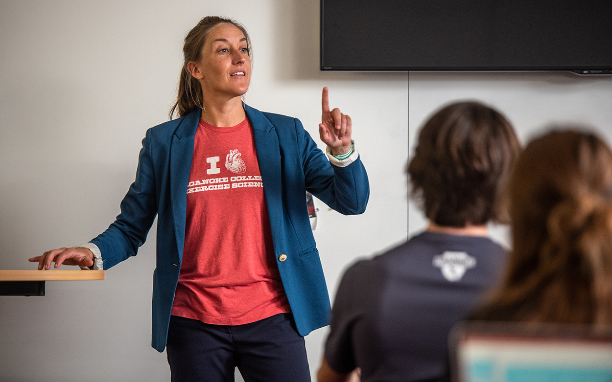 Liz Ackley, wearing a salmon-colored t-shirt and navy blazer, stands in front of a class and holds up one finger as she makes a point during a lecture.