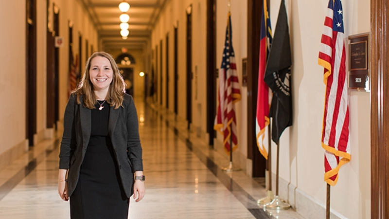 A student dressed in professional attire smiles for a photo in a government building lined with U.S. flags.