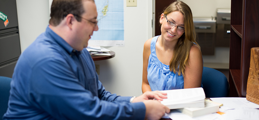 Professor and student sit at a desk looking at a book.