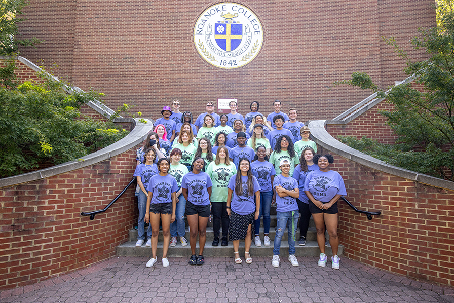 A group of students in identical purple t-shirts stand on the stairs in front of a brick wall that bears the Roanoke College seal, in blue and yellow on a white background.