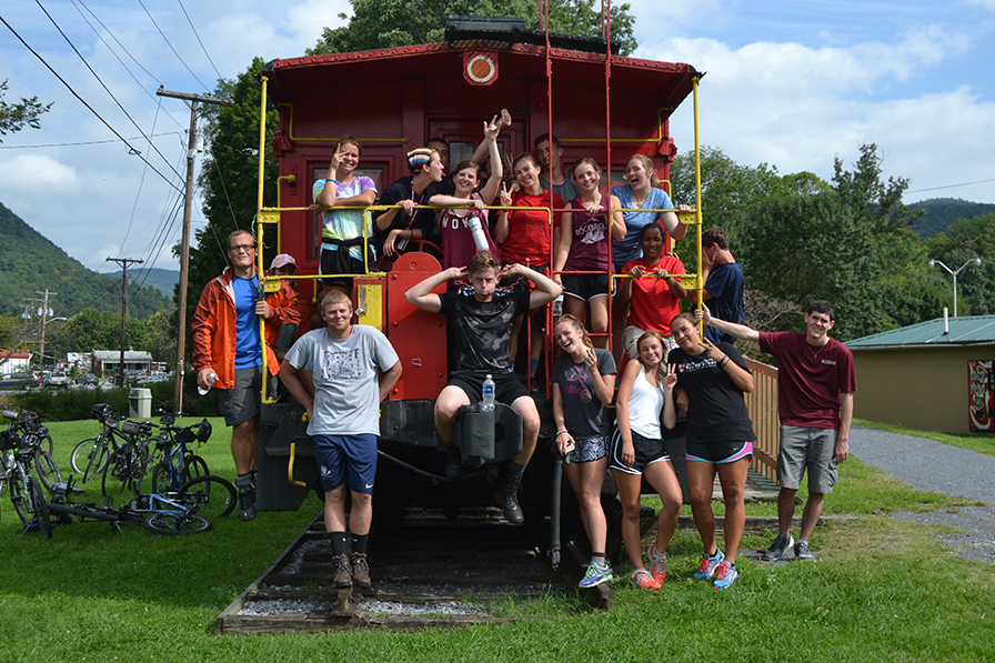 A group of students gather around the back of an old red caboose, with some standing on the ground and others on the caboose platform. They smile and mug for the camera.