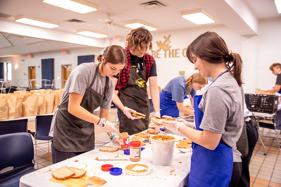 A male student and two female students, all wearing aprons, stand around a table where they are making a bunch of peanut butter and jelly sandwiches for residents of a community shelter.