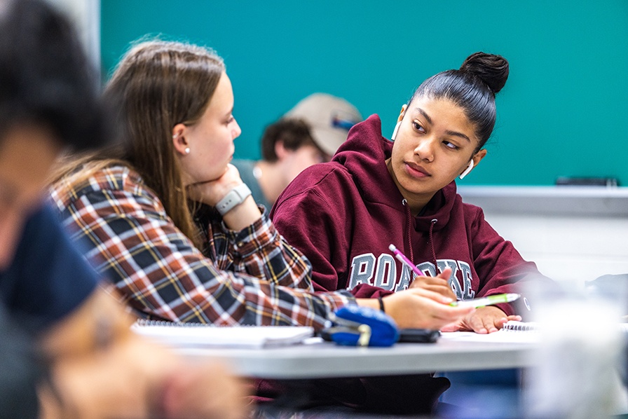 Two female students side by side at desks with a chalkboard behind them