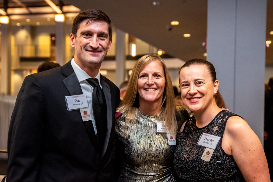 A young male alumni and two female alumni pose together with a photo at an alumni event, where they are dressed up in suits and dresses and wearing name tags.