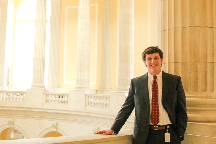 A young man in a dark suit and red tie stands in front of a government building with huge white columns behind him.