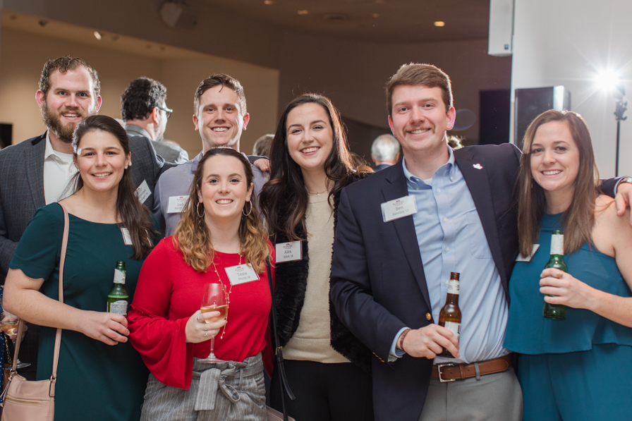 A group of men and women pose for a photo at a networking event, where they are holding cocktails and wearing name tags and smiling.