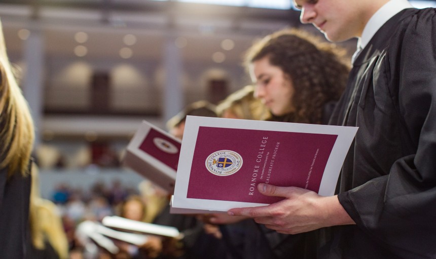 Students in graduation regalia at Baccalaureate reading through the program while seated in the Cregger Center