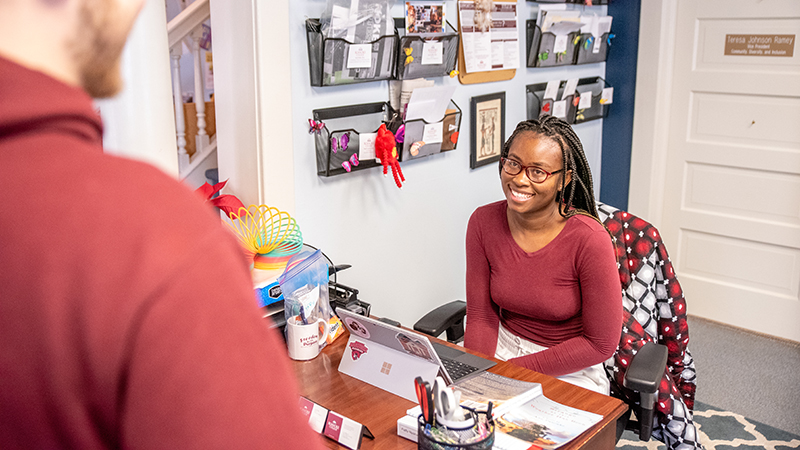 A student sits at a desk in the Chaplain's Office and talks to another student.