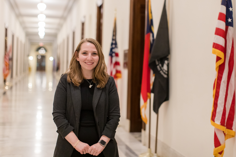 A female student in a dark suit stands in a legislative hallway, and each office behind her has a large flag standing outside of it. 