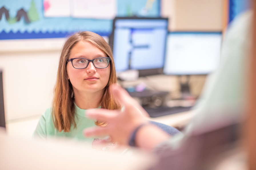 A female student sitting behind a desk in the Residence Life office listens to someone talking to her out  of the camera frame.