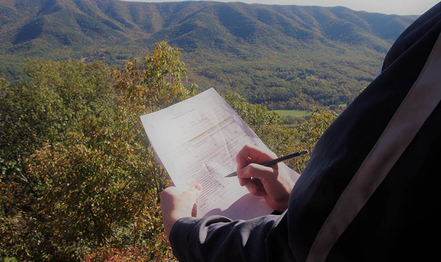 student writing on a sheet while overlooking mountains and a forest