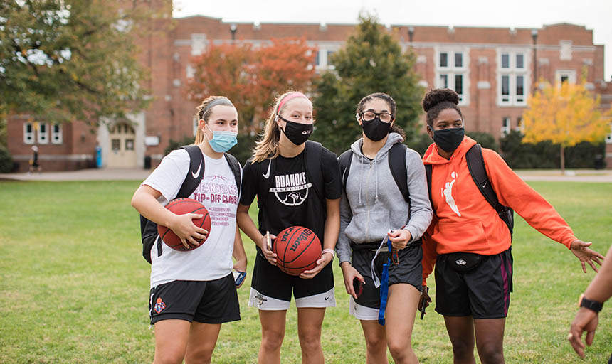 Four female students wearing masks on the back quad. One of them is holding a basketball