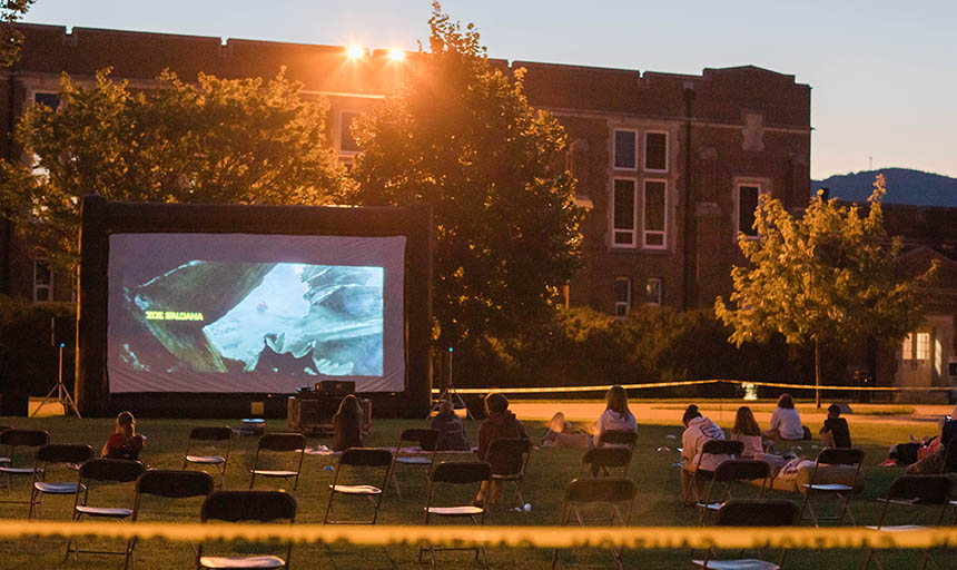 Students sitting in socially-distanced chairs in front of a large movie screen on the back quad