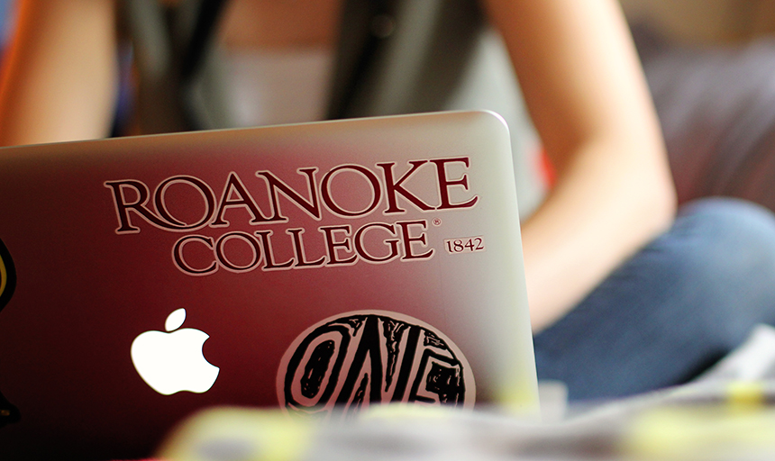 A student sitting at a laptop with a Roanoke College sticker
