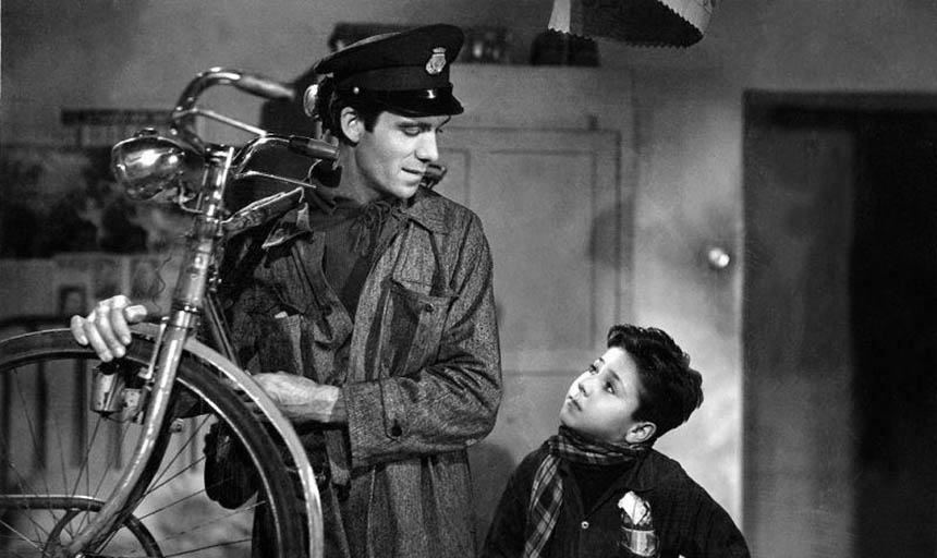 A black and white photo of a man carrying a bike as a child looks up at him