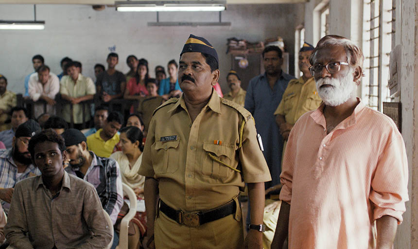 Actors in a scene of Court where two men stand before a room full of seated adults.