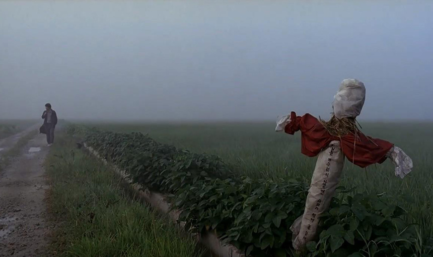 A gloomy image of a field with a scarecrow in the foreground and a lone man walking in the background