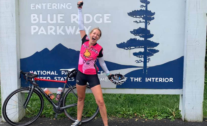 Liz Ackley standing in front of blue ridge parkway sign with her boke and one hand up in the air