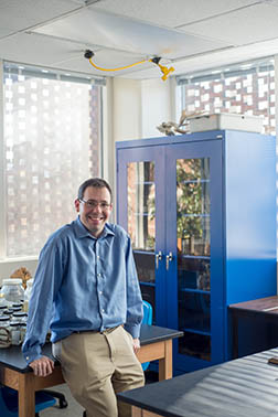 Chris Lassiter leaning against a lab table in his classroom with a see through cabinet and windows behind him