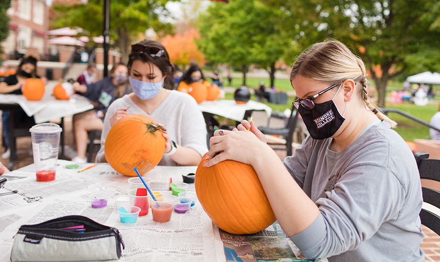 Two students paint pumpkins outdoors while wearing face coverings