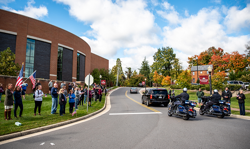 People lining street on campus watching SUV and motorcycles