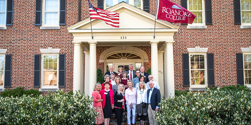 Several people stand in front of brick home with shrubbery on the sides