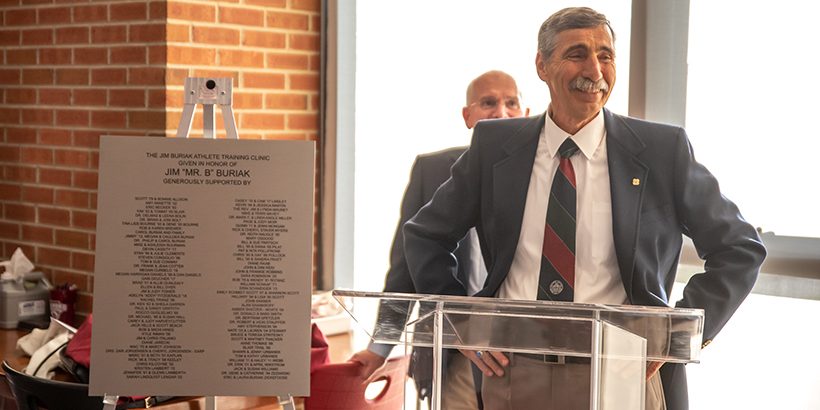 Man stands at podium with his hands on his hips. plaque is to the left of him and another man in background